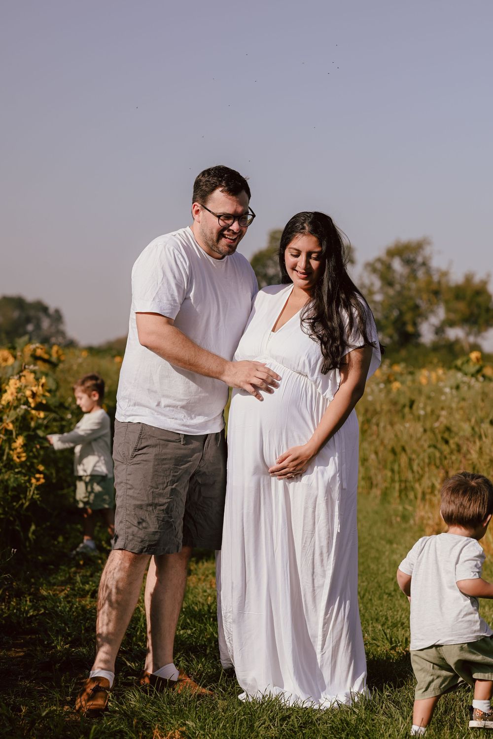 Family in white attire poses together in a sunny sunflower field during golden hour.
