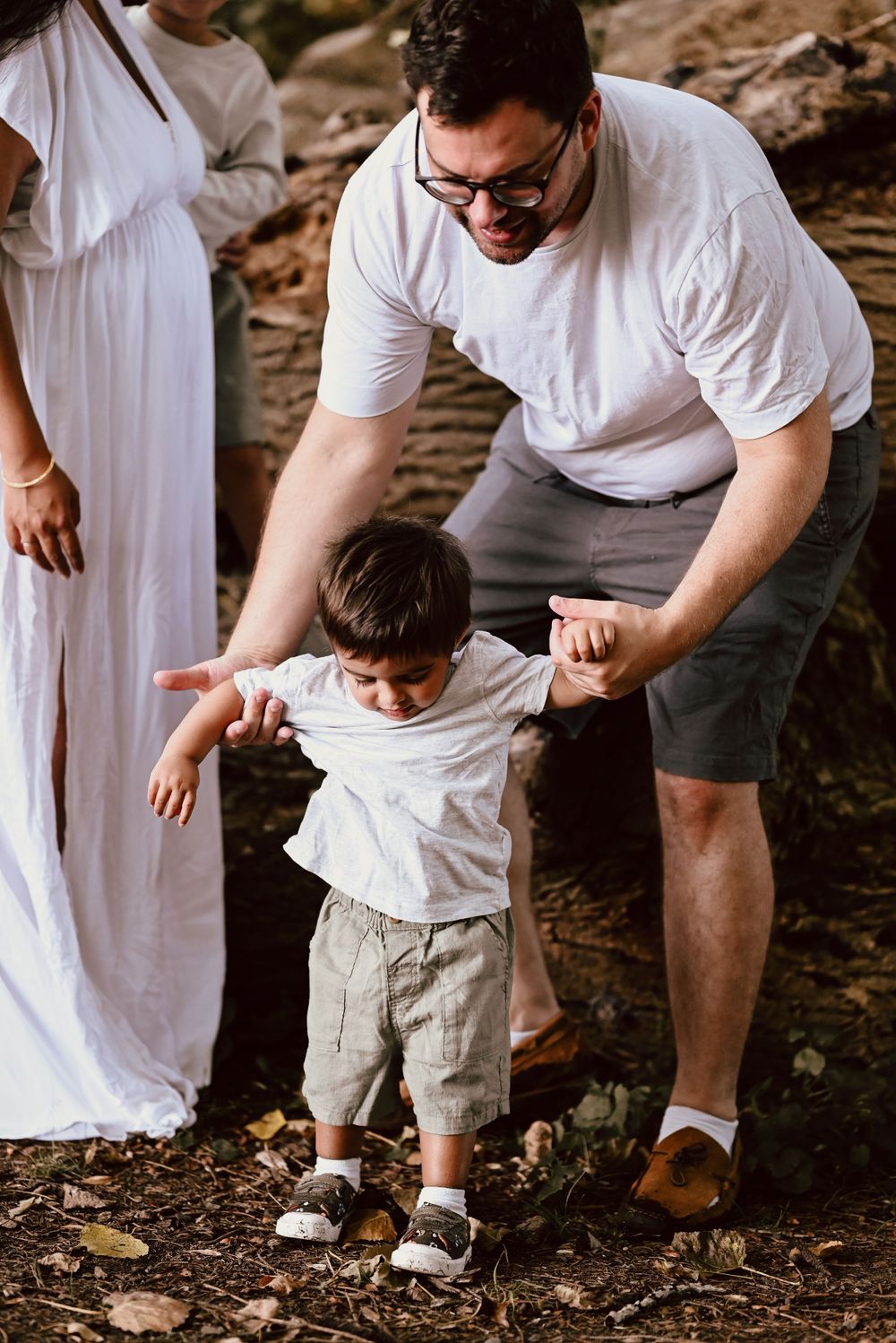 Person in white shirt helps small child balance and walk on rocky terrain while someone in white dress stands nearby.