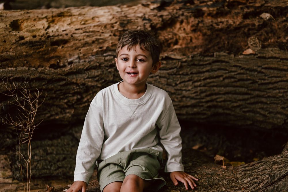 Portrait of a child wearing a white long sleeve shirt and khaki shorts sitting by a large wooden log in a rustic outdoor setting.