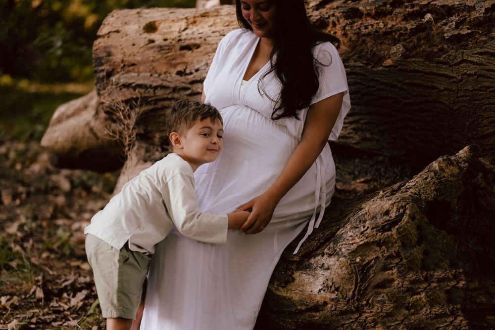 Tender maternity family moment captured outdoors against natural textured log backdrop in warm sunlight.