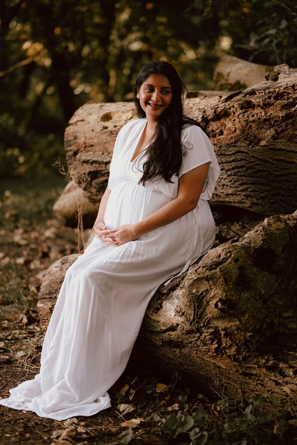 A person in a flowing white dress sits on fallen logs in a wooded setting during a maternity photo session.