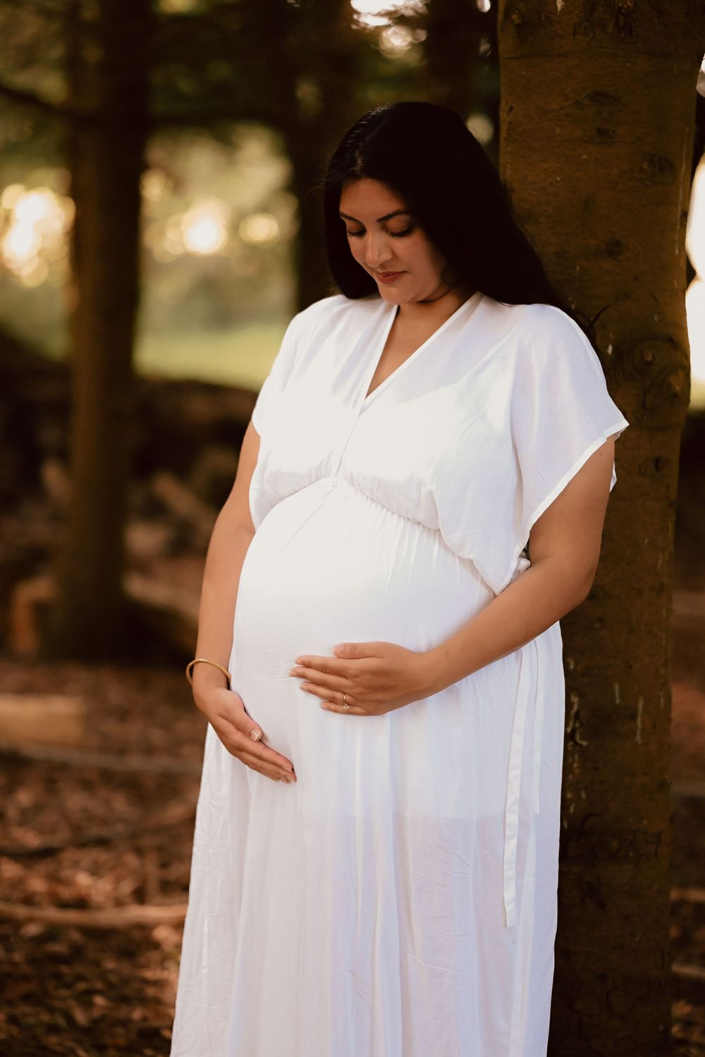 An expectant mother in a flowing white dress stands beneath a tree in a dreamy outdoor maternity photoshoot.