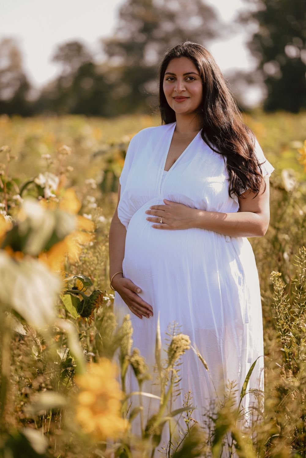 A pregnant woman in a white dress stands among wildflowers in a sunlit meadow during golden hour.