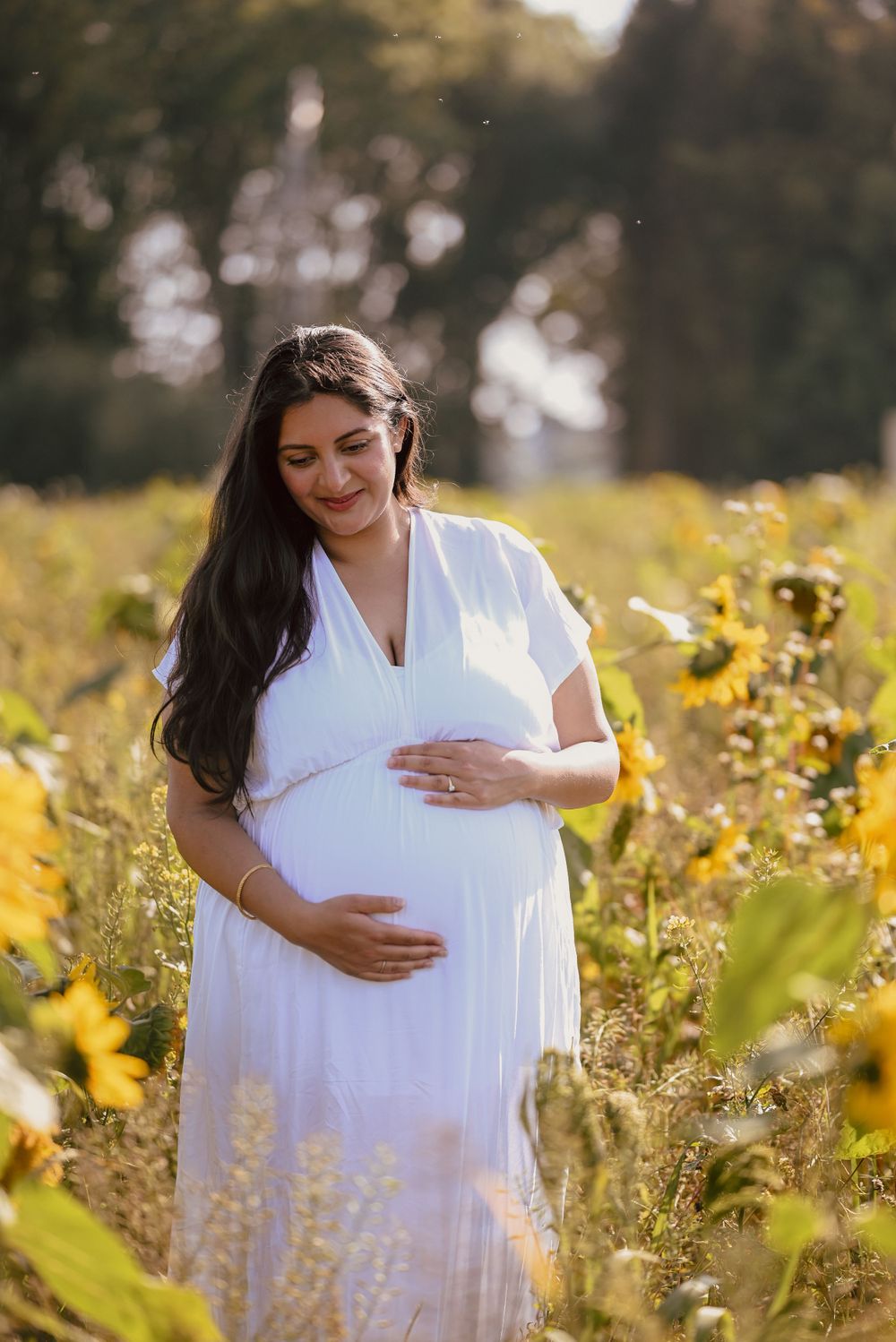 A pregnant woman in a flowing white dress stands among yellow sunflowers in a sunlit meadow.