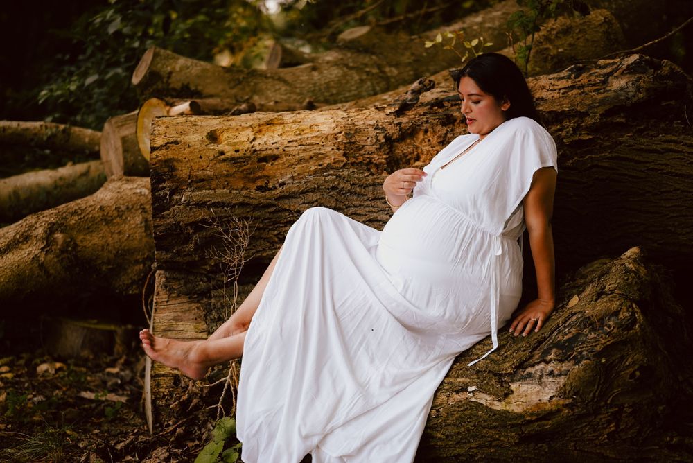 A pregnant woman in a flowing white dress poses serenely on a fallen log in a peaceful forest setting.
