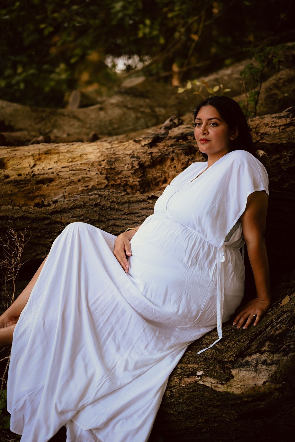 Maternity portrait in white flowing dress against natural wood backdrop with moody forest lighting.