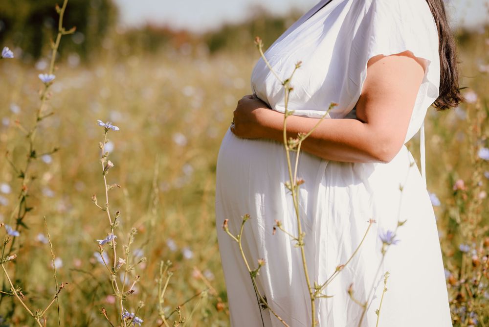 Pregnant Person in a flowing white dress stands in a sunlit meadow surrounded by delicate wildflowers and tall grass.