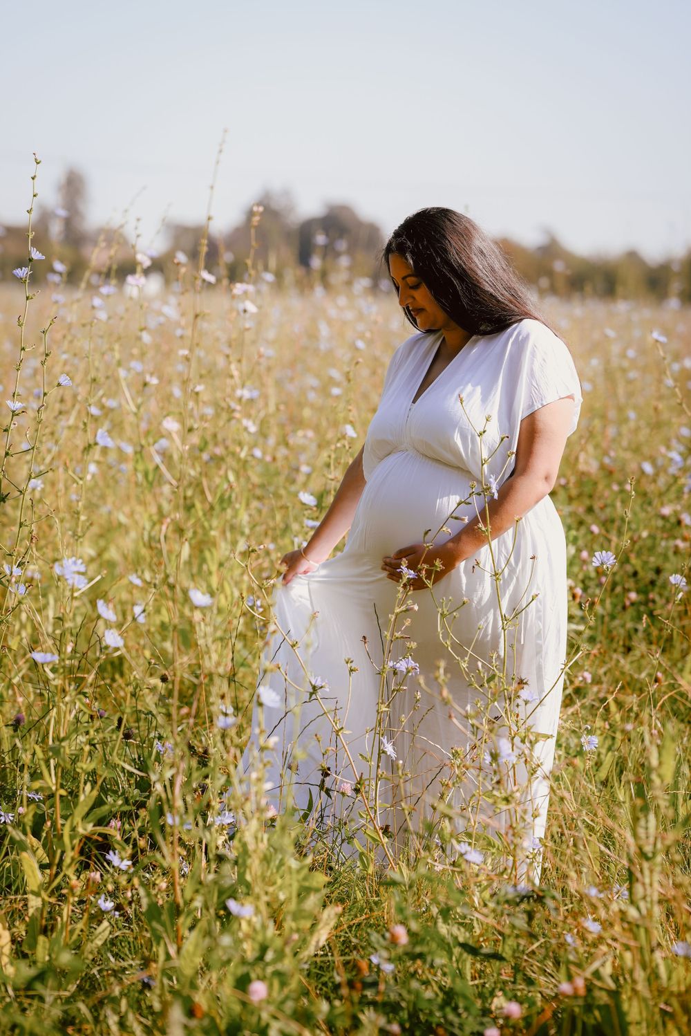 Expectant mother in flowing white dress standing peacefully in a summer wildflower meadow during golden hour.
