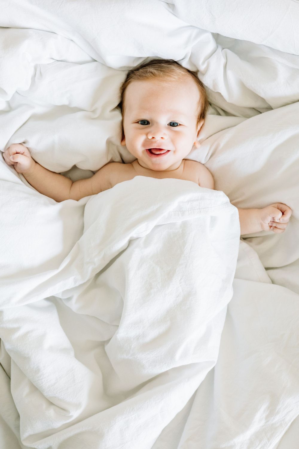 A happy infant lying in white bedding and smiling at the camera.