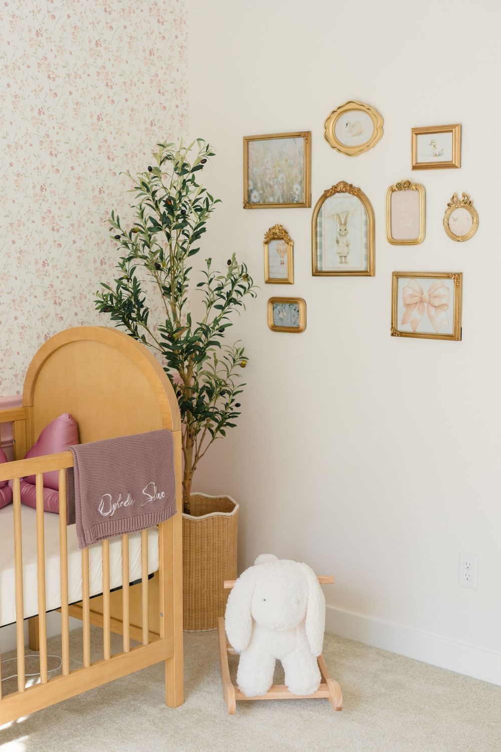 Cozy nursery with wooden crib, floral wallpaper, gold gallery wall frames, plush bunny rocker, and potted olive tree.
