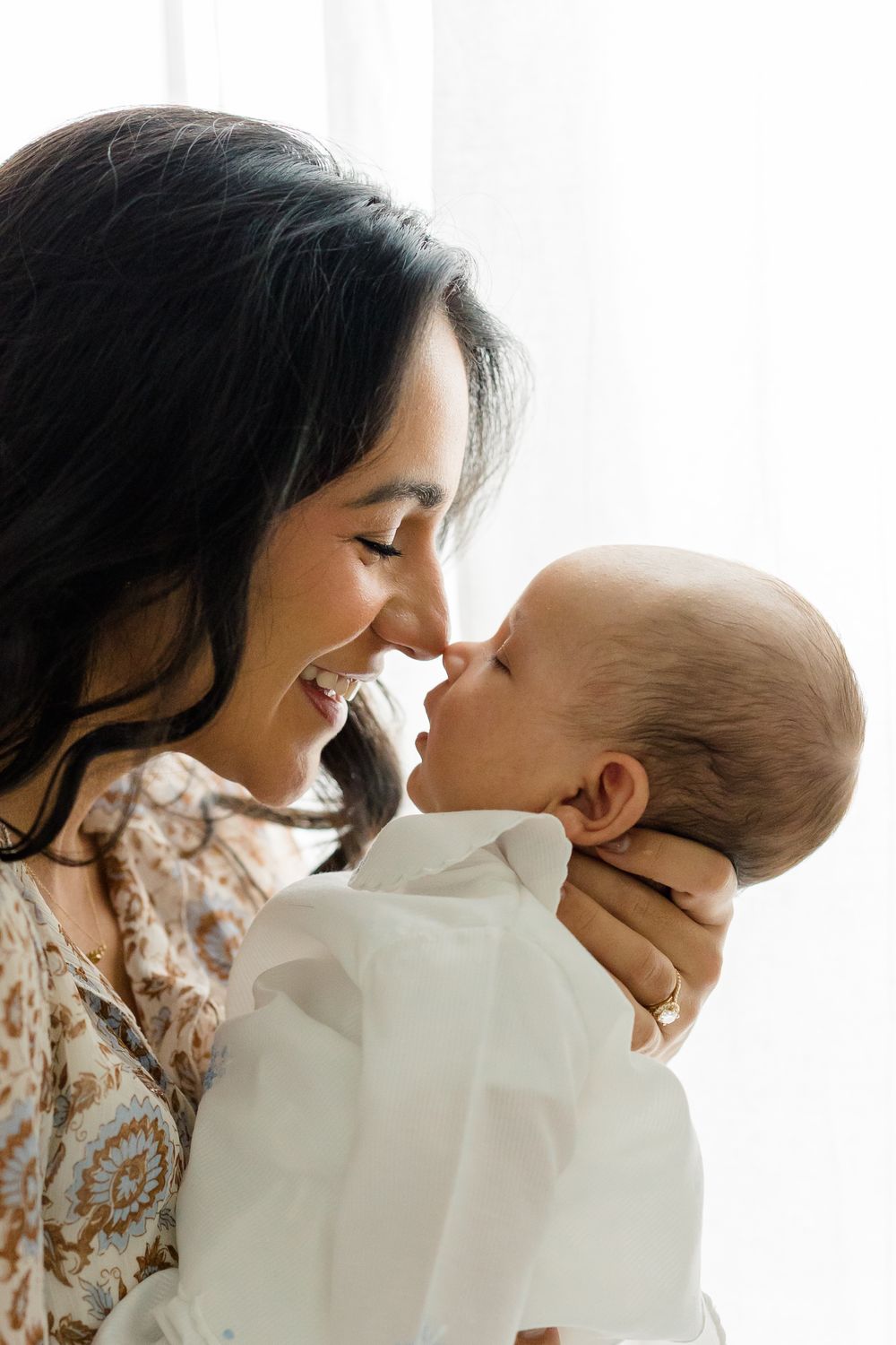 A smiling dark-haired mother tenderly touches noses with her newborn baby wrapped in white cloth.