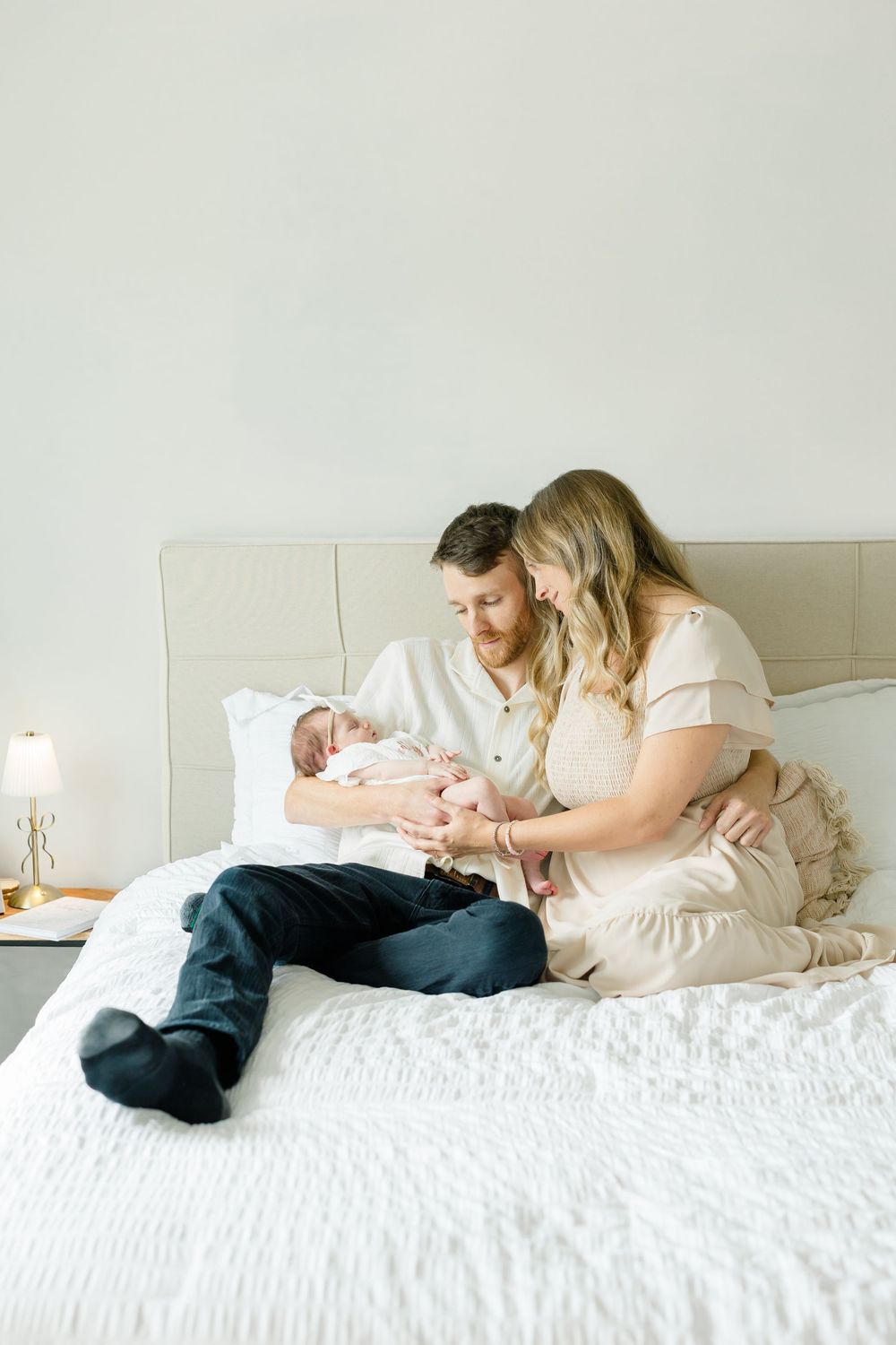 A couple sits on a white bed, lovingly gazing at their newborn baby in a bright, softly lit bedroom.