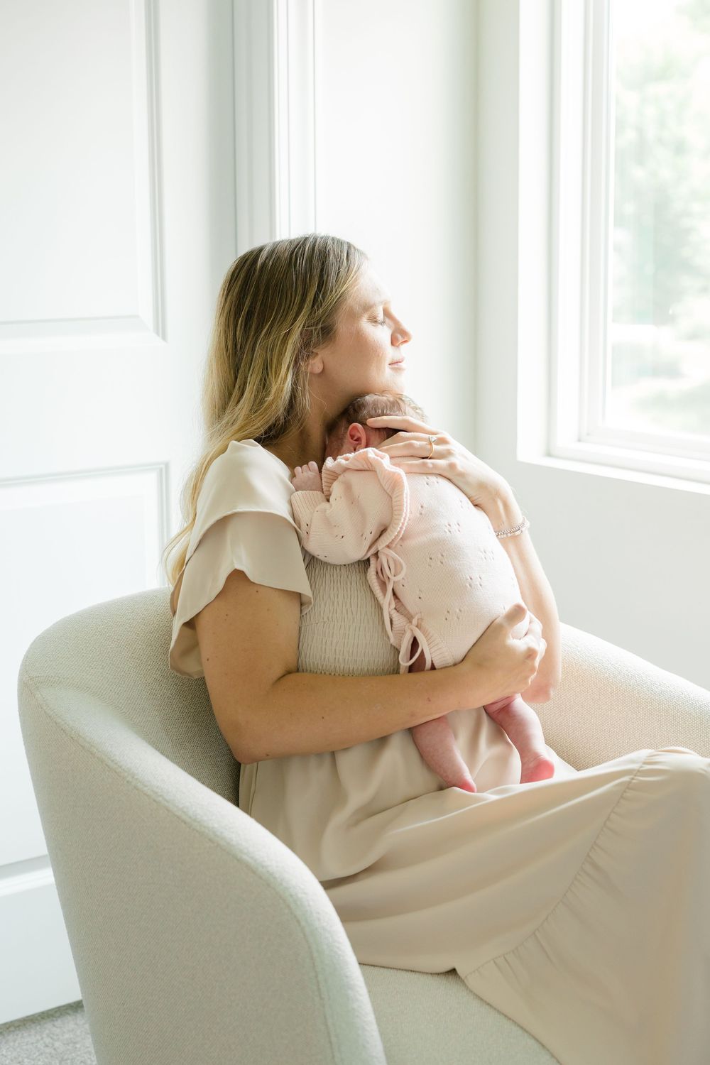A mother with eyes closed holds her newborn baby in a cream chair by a bright window, both dressed in soft pink and beige.