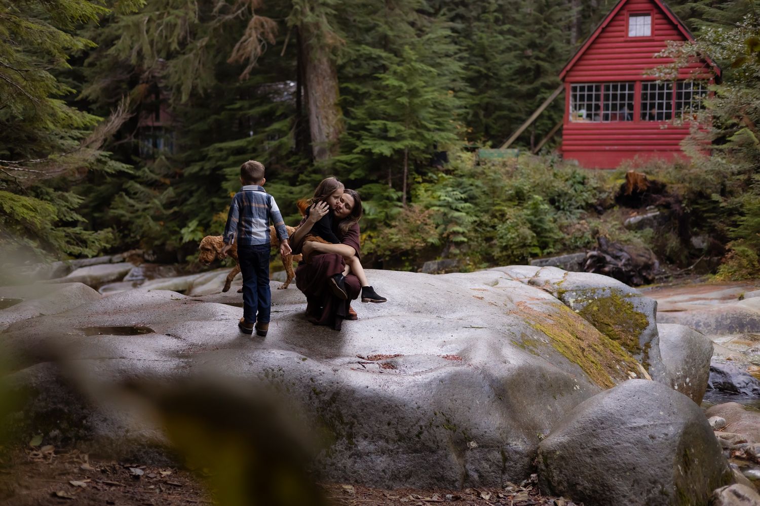 A mom with her kids and their dog play on a large rock in front of a red cabin during a family adventure photo session in Washington