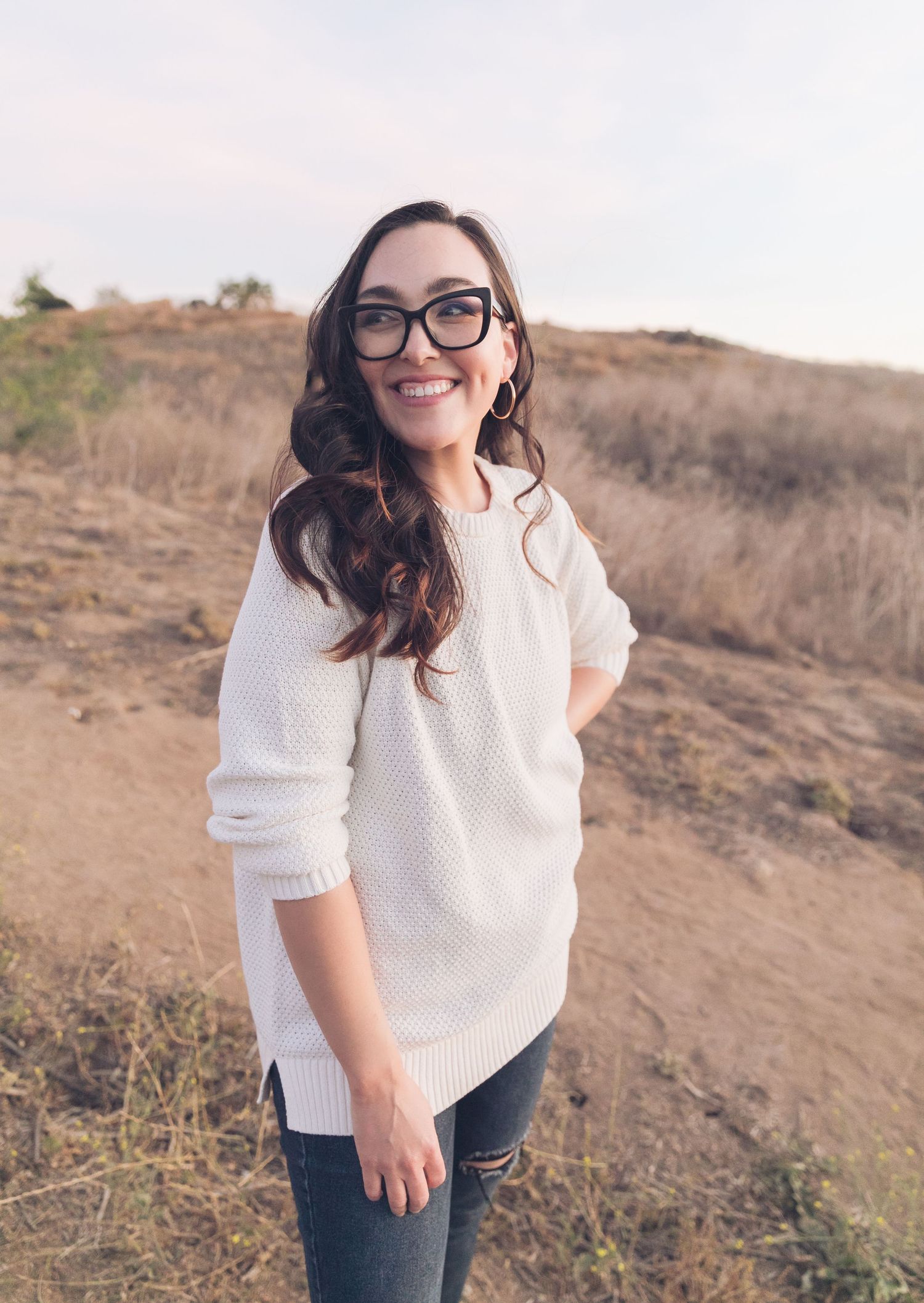 Portrait of person in white sweater and glasses smiling outdoors at sunset.