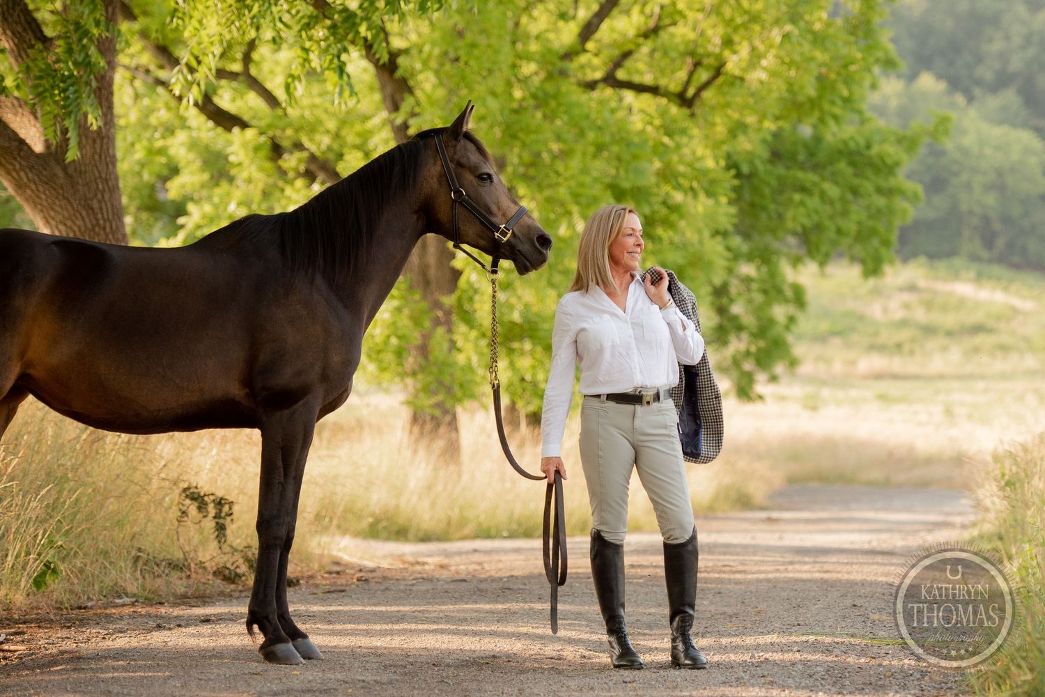 Equestrian in riding attire stands with a dark horse on a tree-lined path in natural sunlight.