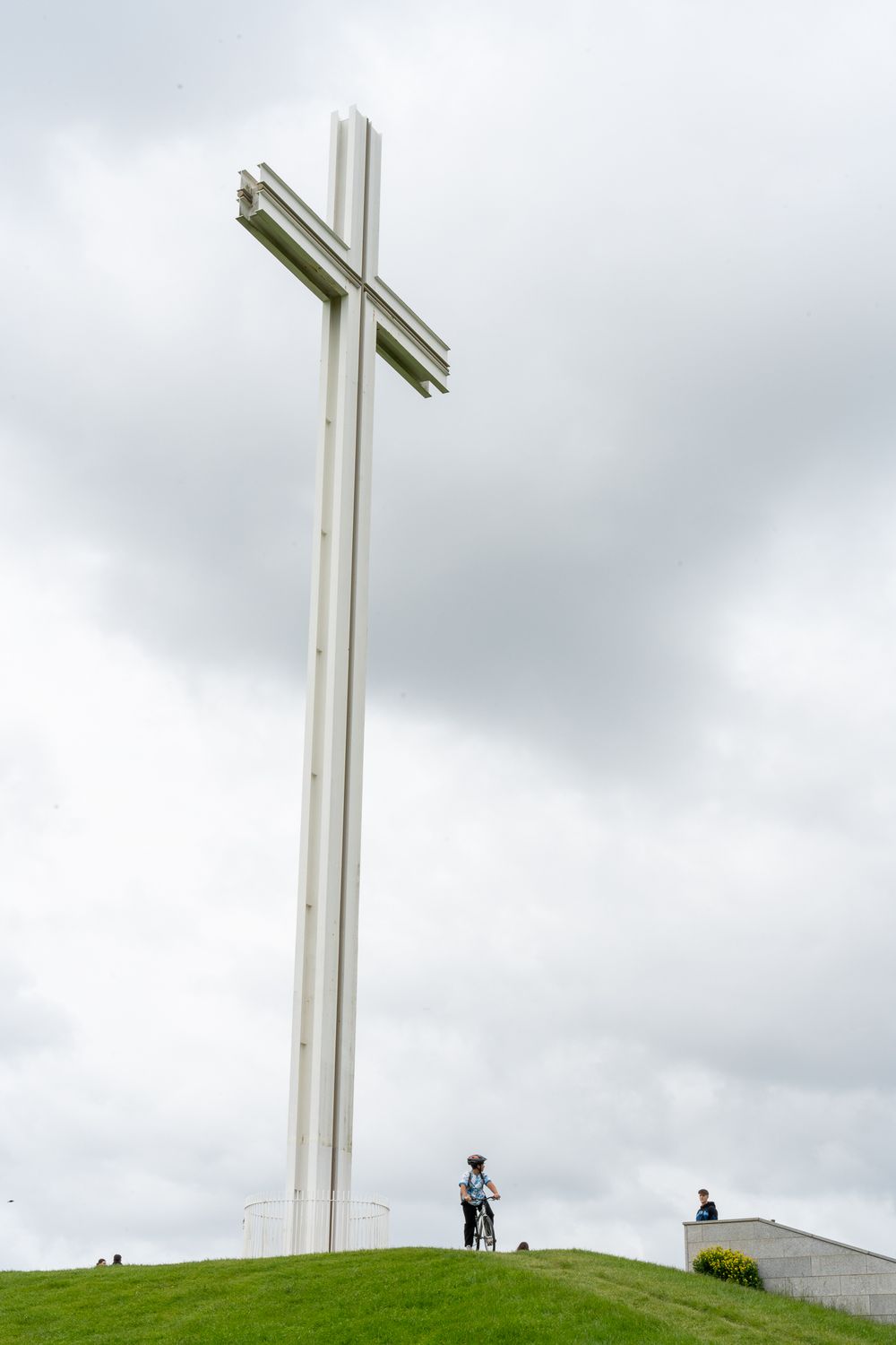 A tall white cross stands against cloudy skies on a grassy hilltop.