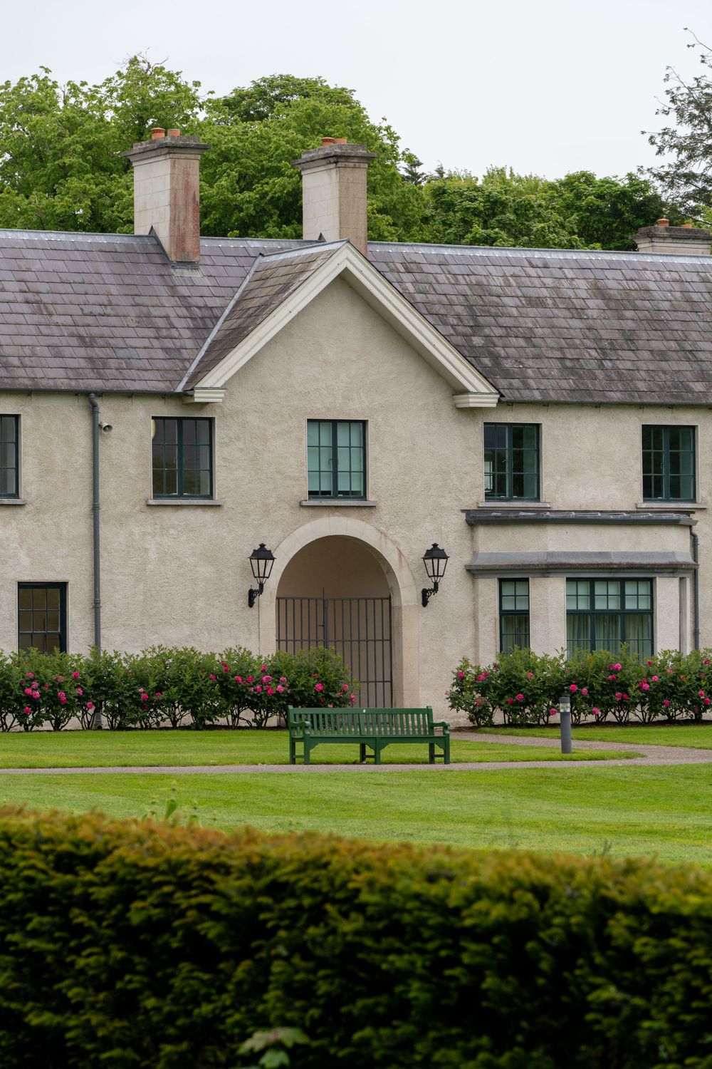 A grey stone house with twin chimneys and an arched entrance, featuring landscaped gardens and a bench out front.