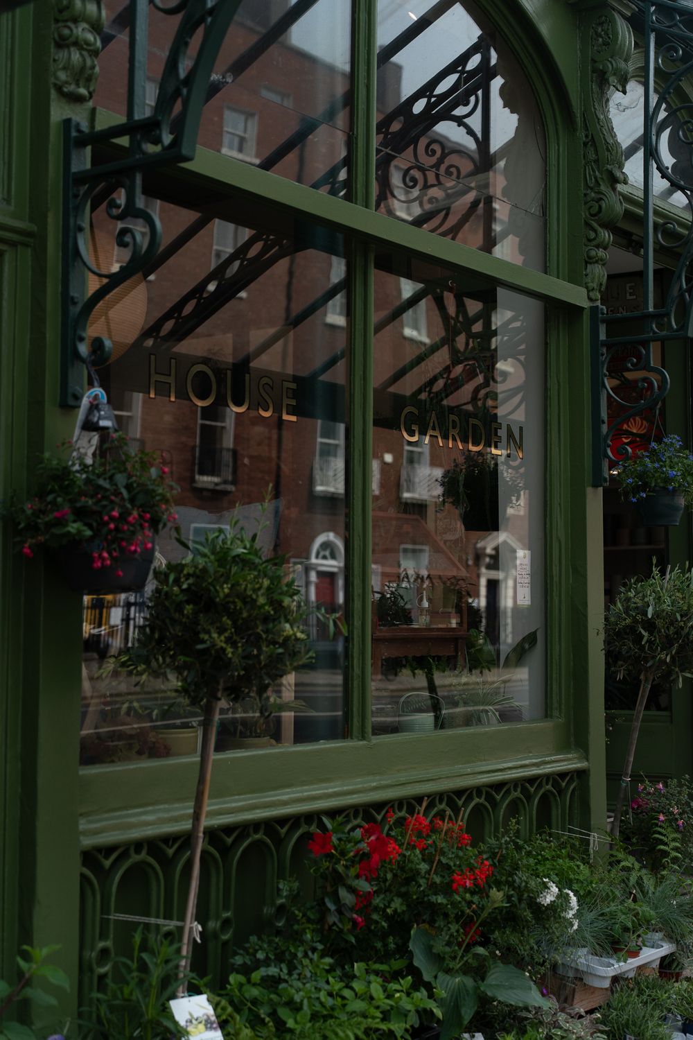 Ornate green ironwork window frame with 'House Garden' text and hanging flower baskets.