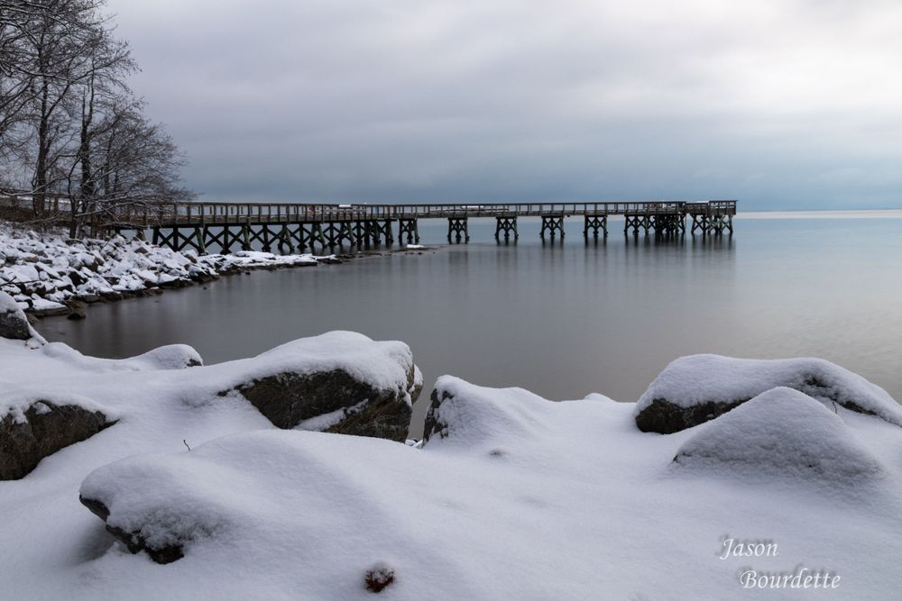 A serene winter bay landscape covered in fresh snow. The calm, icy-blue water reflects the soft light of an overcast sky. Chesapeake Bay
