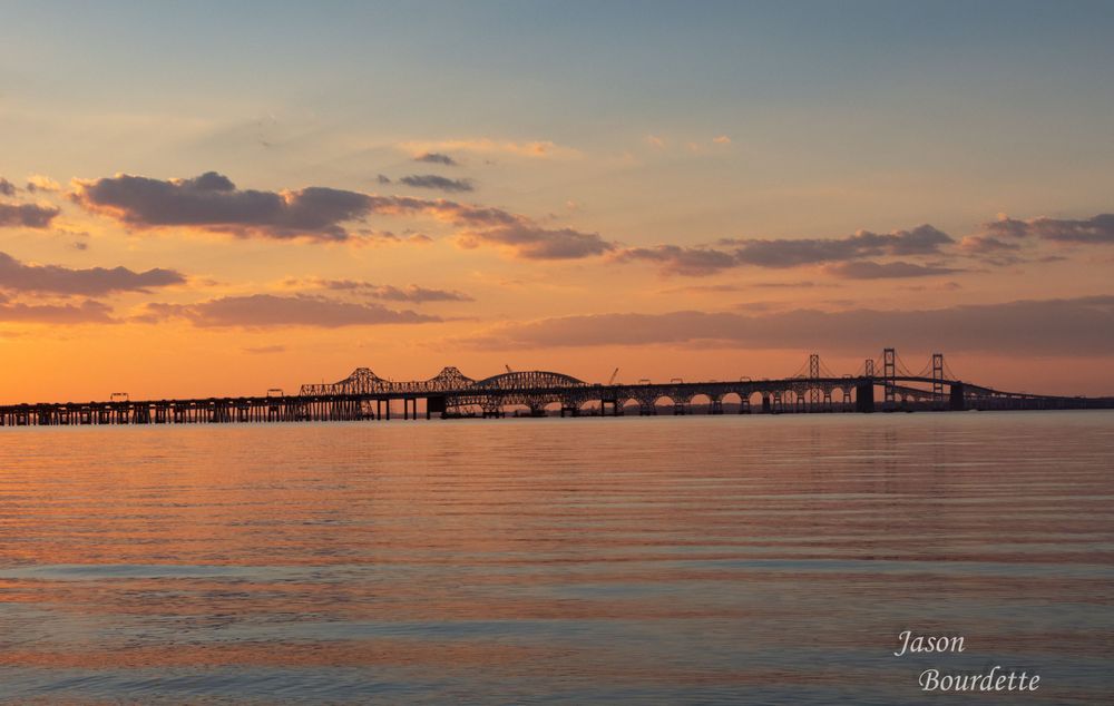 The Chesapeake Bay bridge at sunset stretching in the distance.  The sky is painted with hues of orange and blue, with scattered clouds
