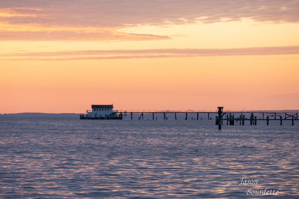 A wooden boathouse extending into the bay at sunrise, the sky glows with warm hues of orange