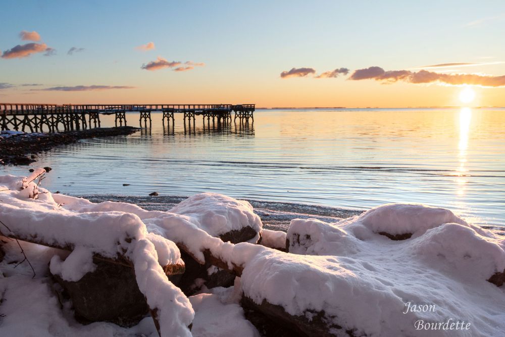 A serene bay with fishing pier is dusted with a layer of fresh snow, creating a striking contrast between the white snow and the sunrise.