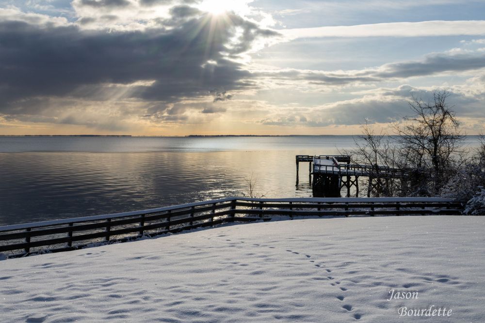 A serene bay with fishing pier is dusted with a layer of fresh snow, creating a striking contrast between the white snow and the sunrise.