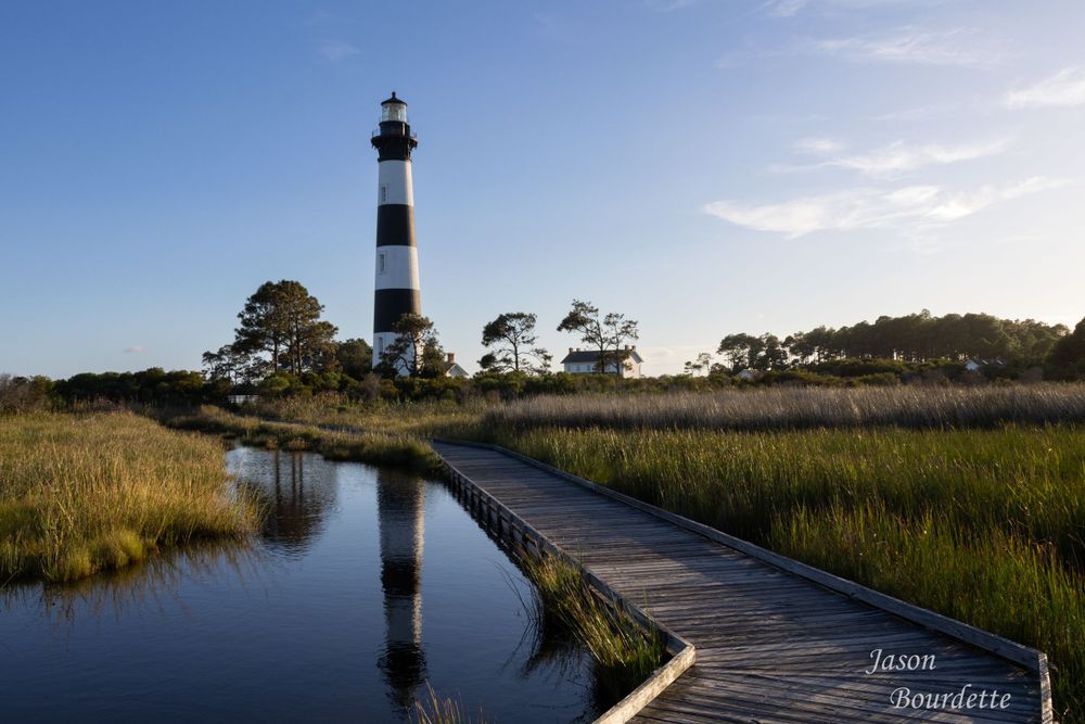 Bodie Island Lighthouse towers against a clear blue sky its black and white stripes standing out amidst the surrounding green marshland land