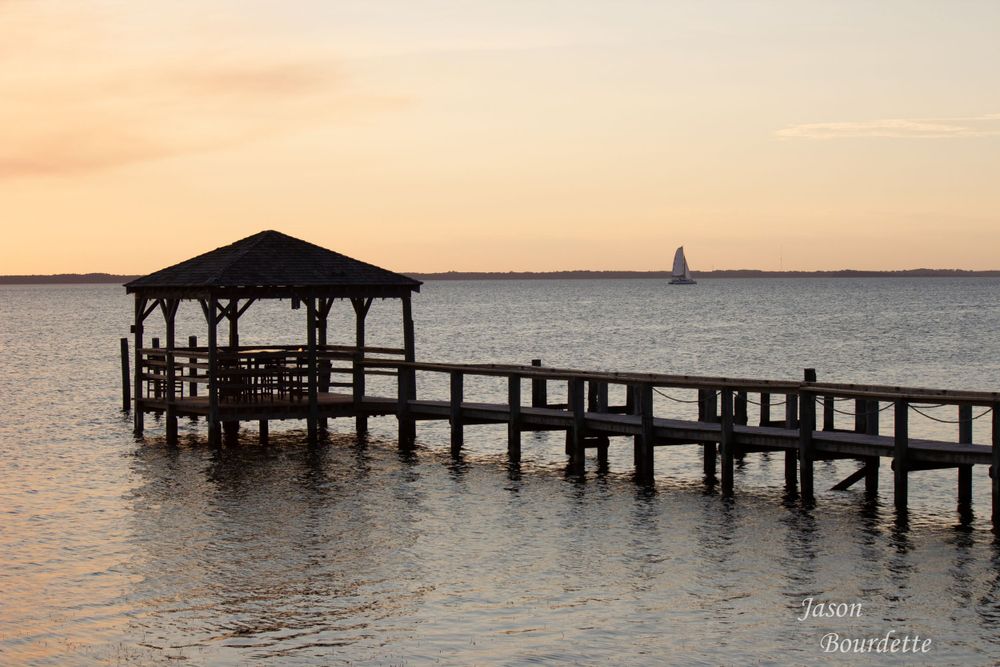 Wooden pier extending into the Sound water at Outer Banks on a calm evening. A sailboat passes by in the distance.