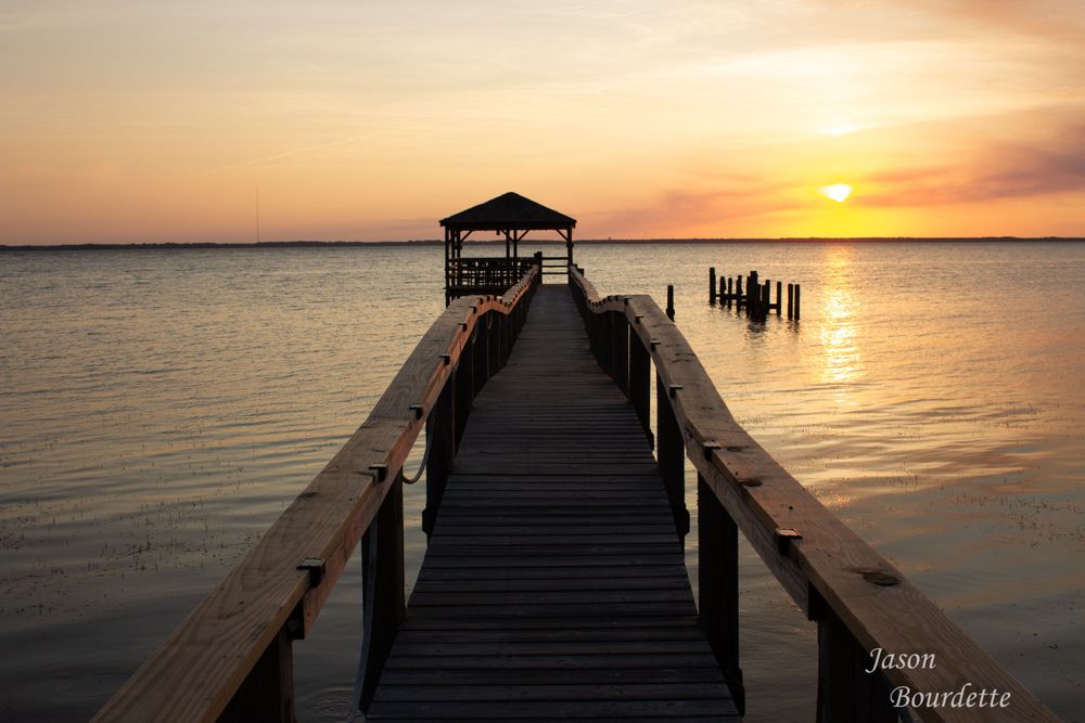 A wooden pier extends out into calm water at sunset, with the sky displaying warm hues of orange, pink, and purple