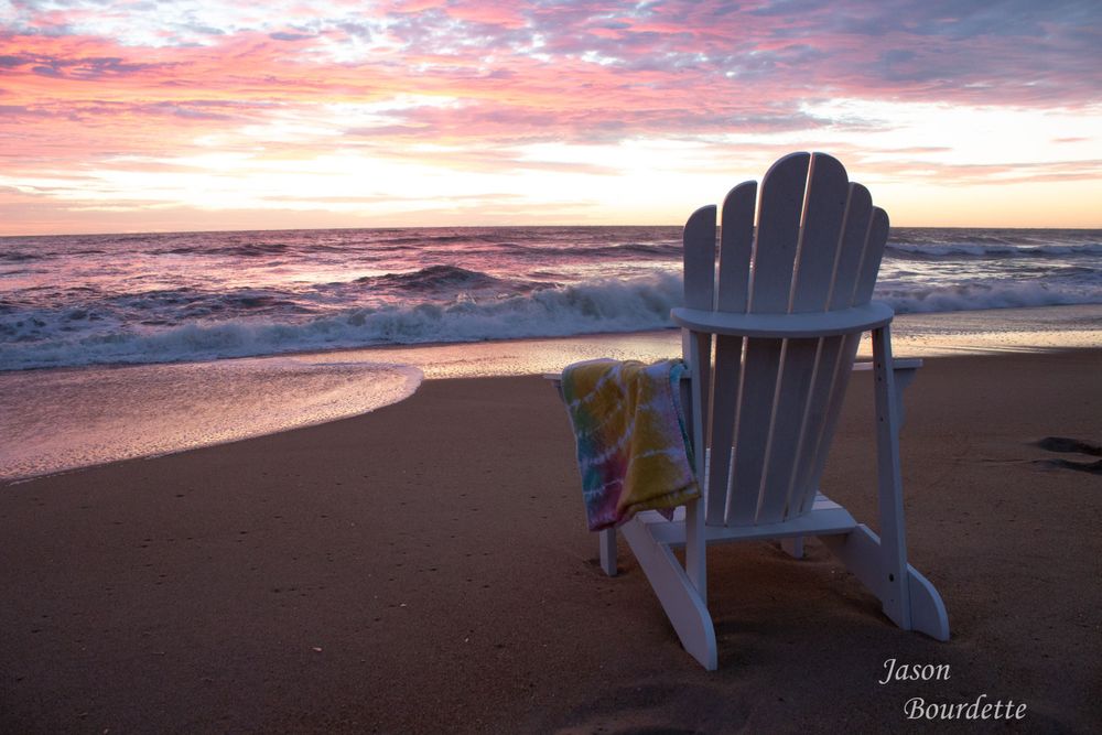An Adirondack chair sits peacefully on the sandy beach at sunrise, facing the gentle waves rolling onto the shore, bathed in morning glow.