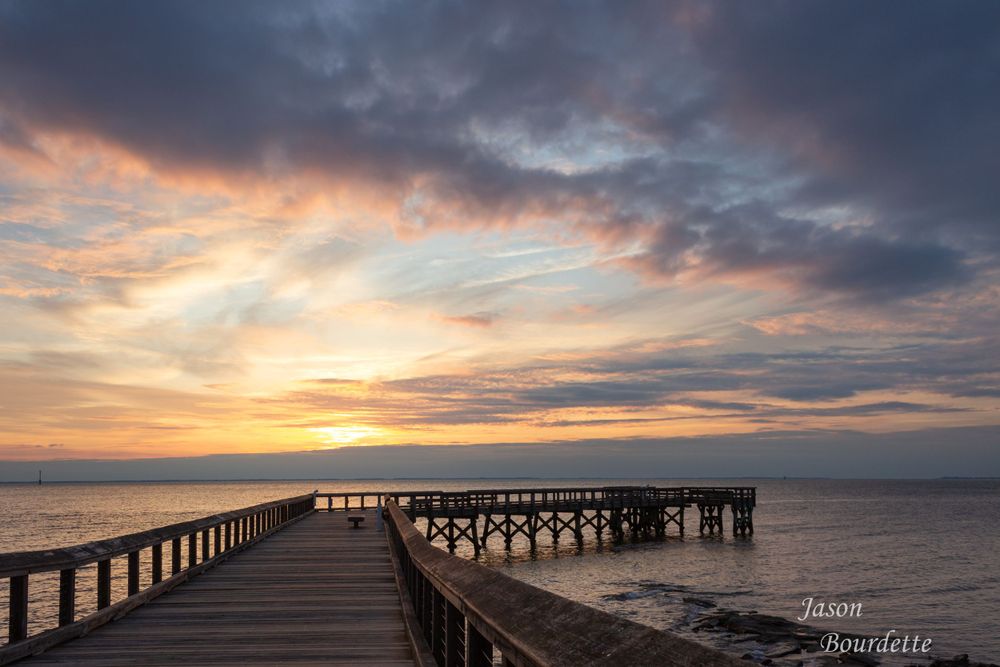 A wooden pier extends out into calm water at sunrise on the Chesapeake Bay. Taken at Downs Park. sky is full of clouds