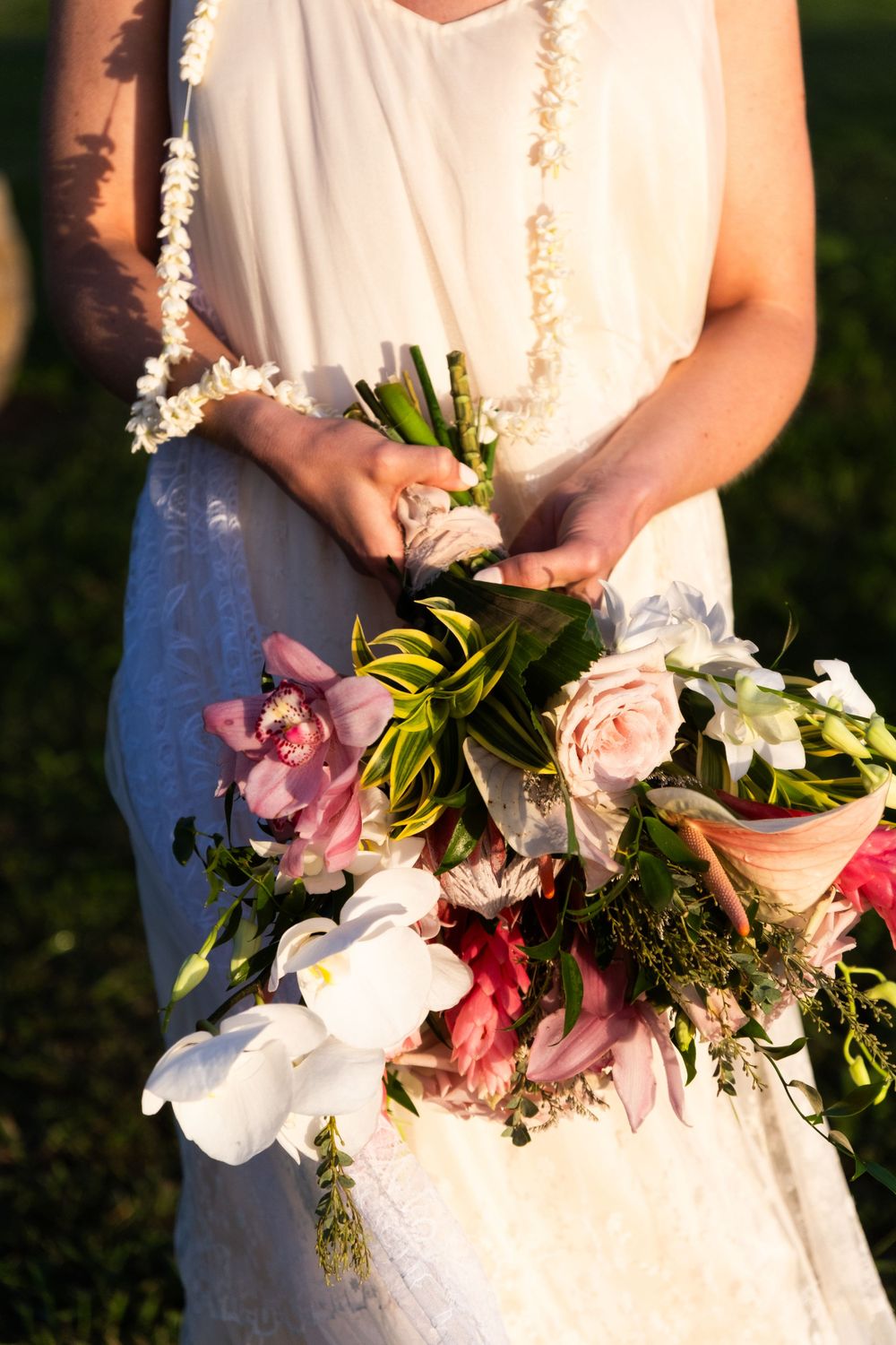 Delicate bridal bouquet featuring pink orchids, white roses, and greenery against a white dress in natural sunlight.