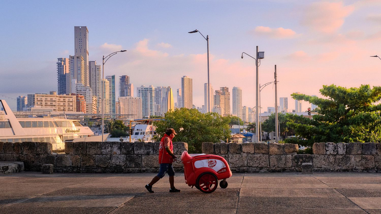 Urban skyline at sunset with person pushing cart along waterfront pathway.