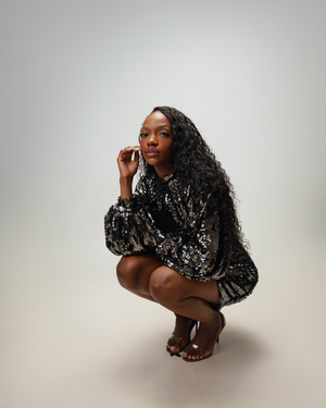 Model crouches in a glittering black sequined dress against a light background.