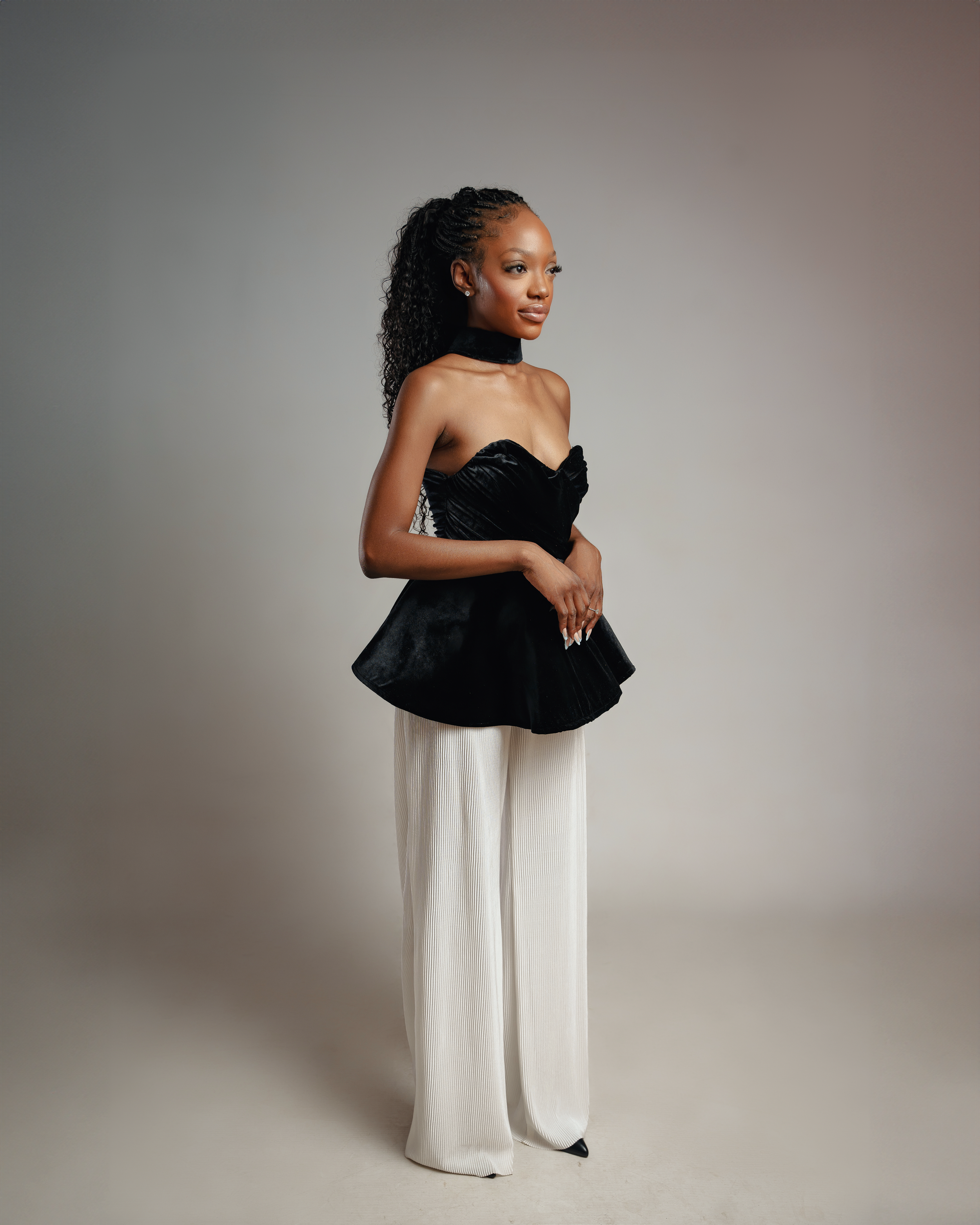 Fashion model in black velvet peplum top and white wide-leg pants poses in studio.