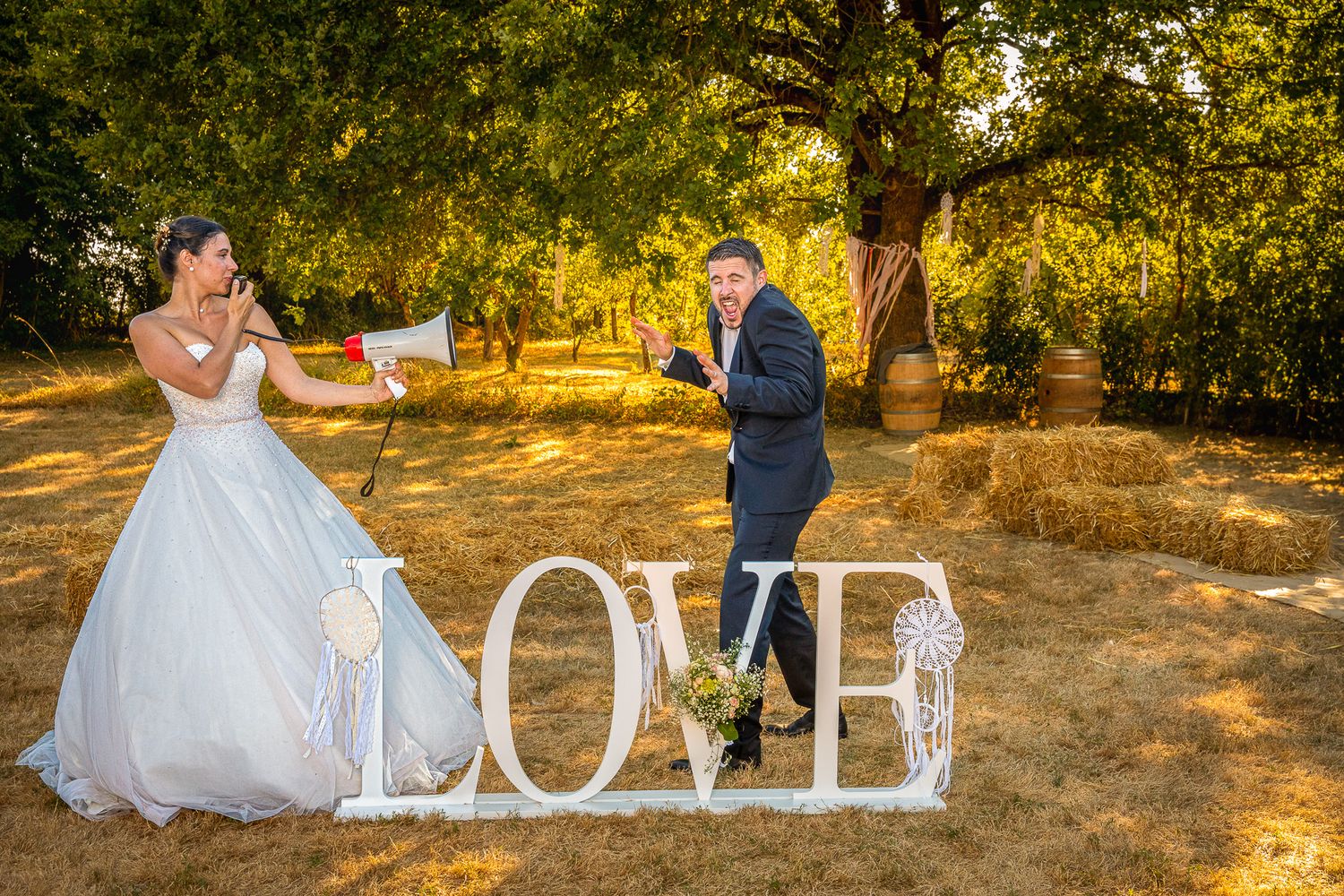 Newlyweds pose playfully with white 'LOVE' letters in an outdoor setting at sunset.