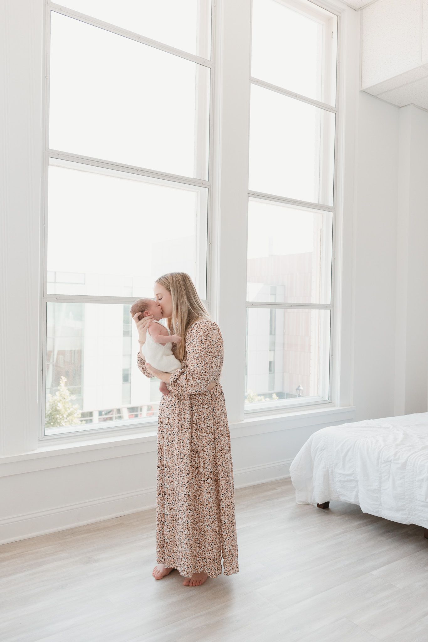 Mother holding her young child near a large window in a bright, minimalist studio, sharing a quiet moment together.