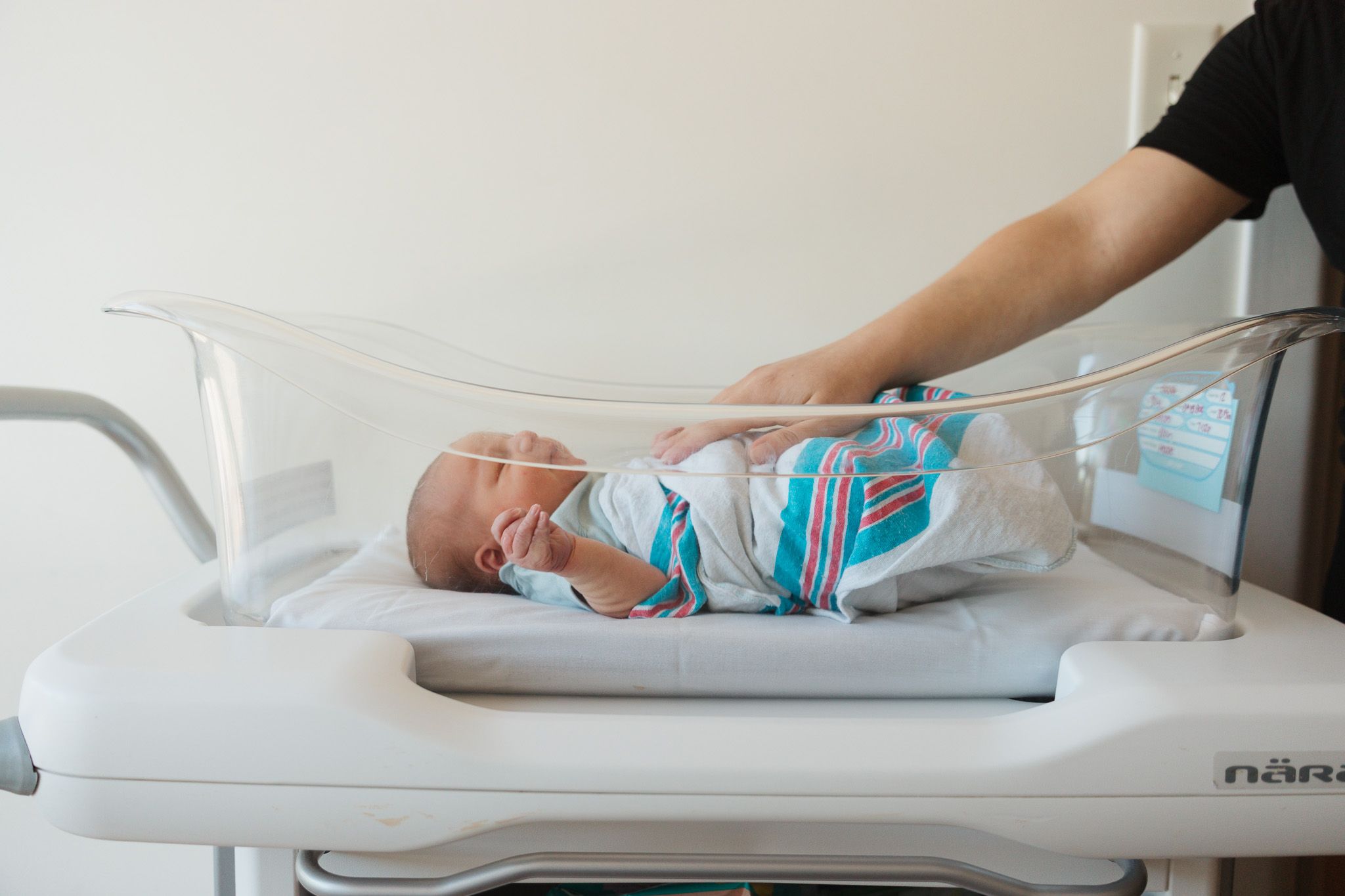 A newborn baby is laying in a clear hospital bassinet while their parent's hand is resting on the babies chest.