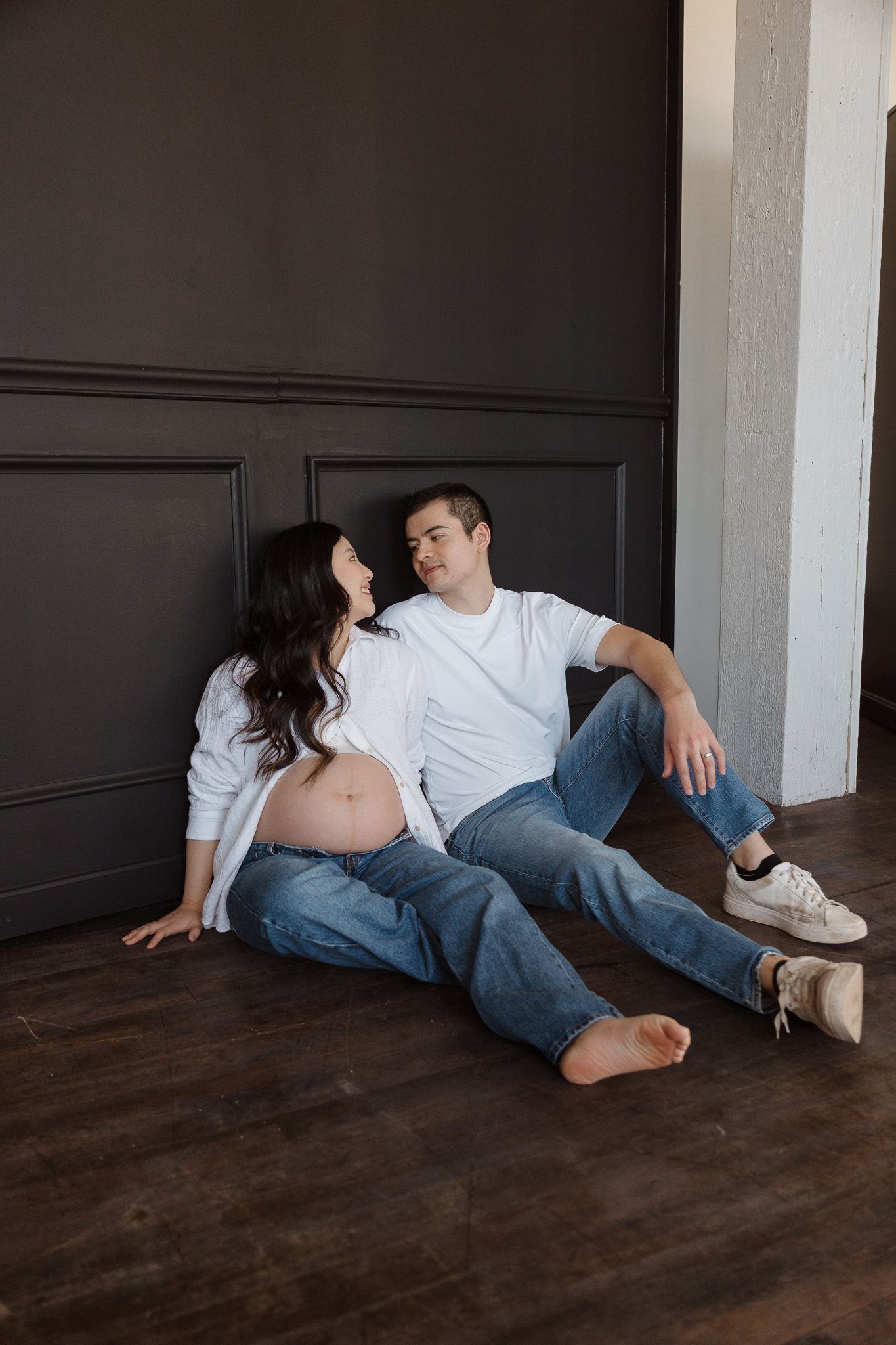 Casual portrait of a couple sitting on the floor against a dark wall, relaxed and smiling together.