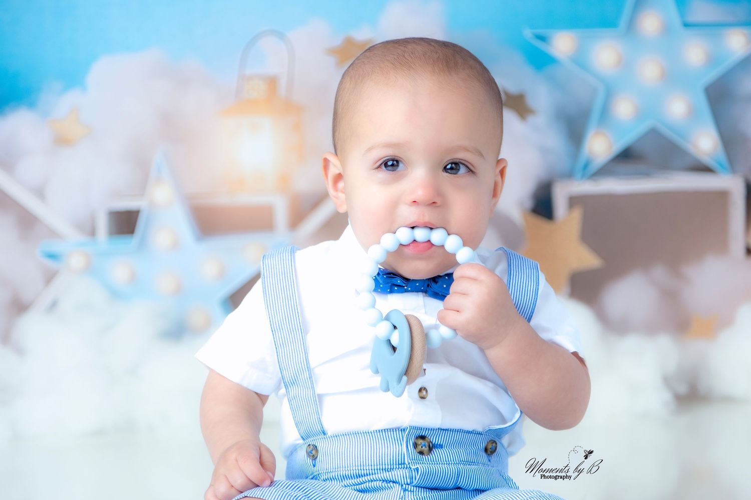 Baby in blue overalls and white shirt playing with toy against cloudy backdrop with stars.