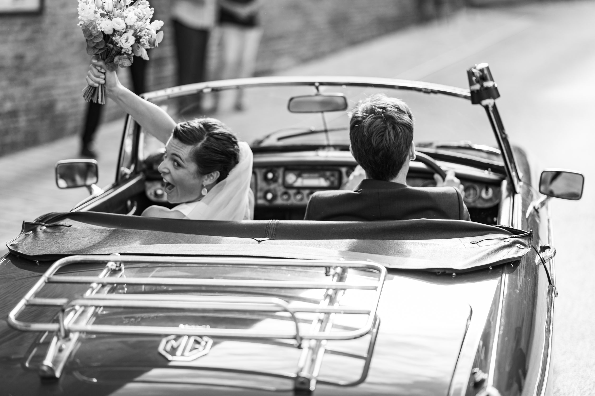 A newlywed couple celebrates in a vintage convertible car, with the bride holding her bouquet up.