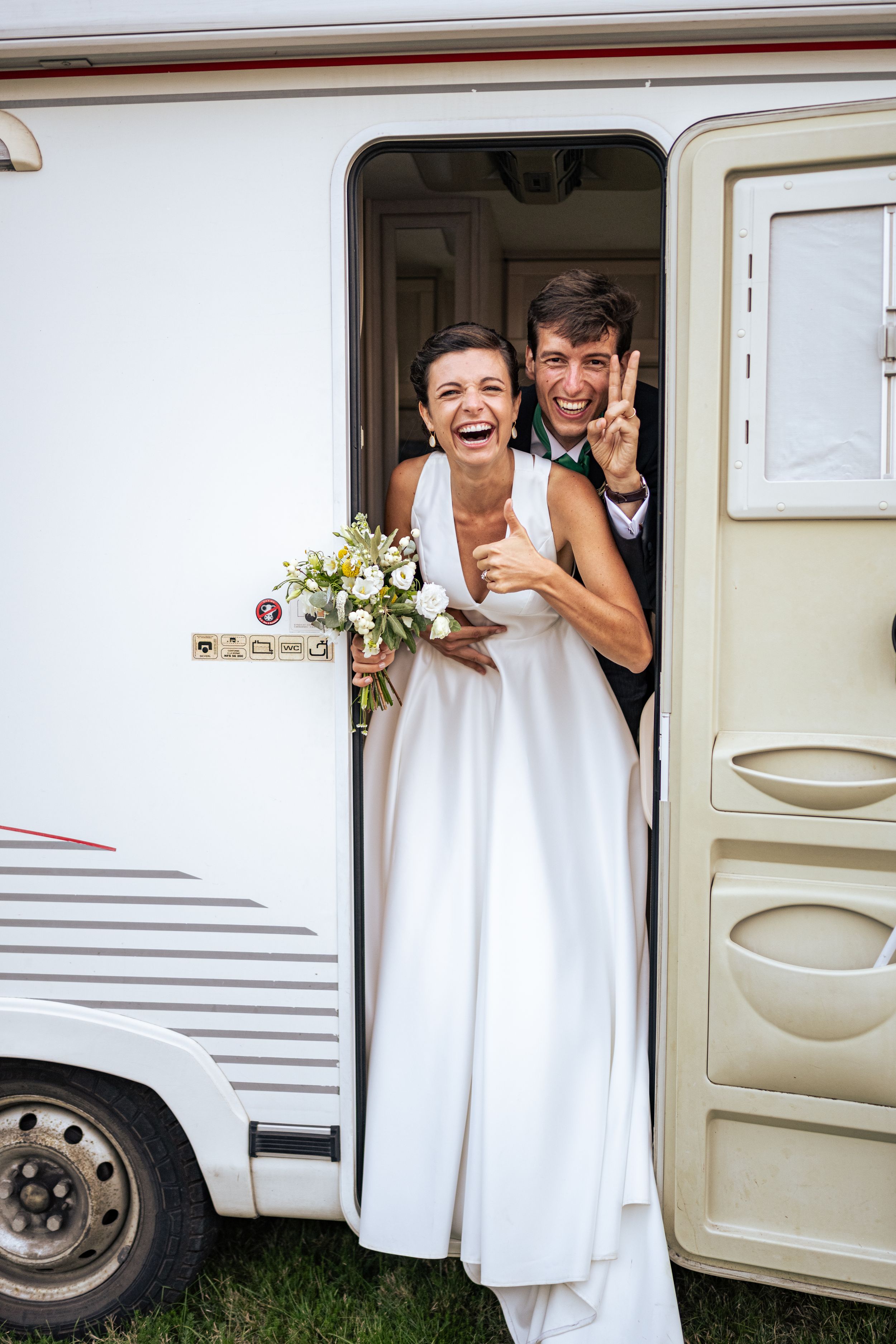 A couple laughs together in the doorway of a white camper van with wedding flowers.