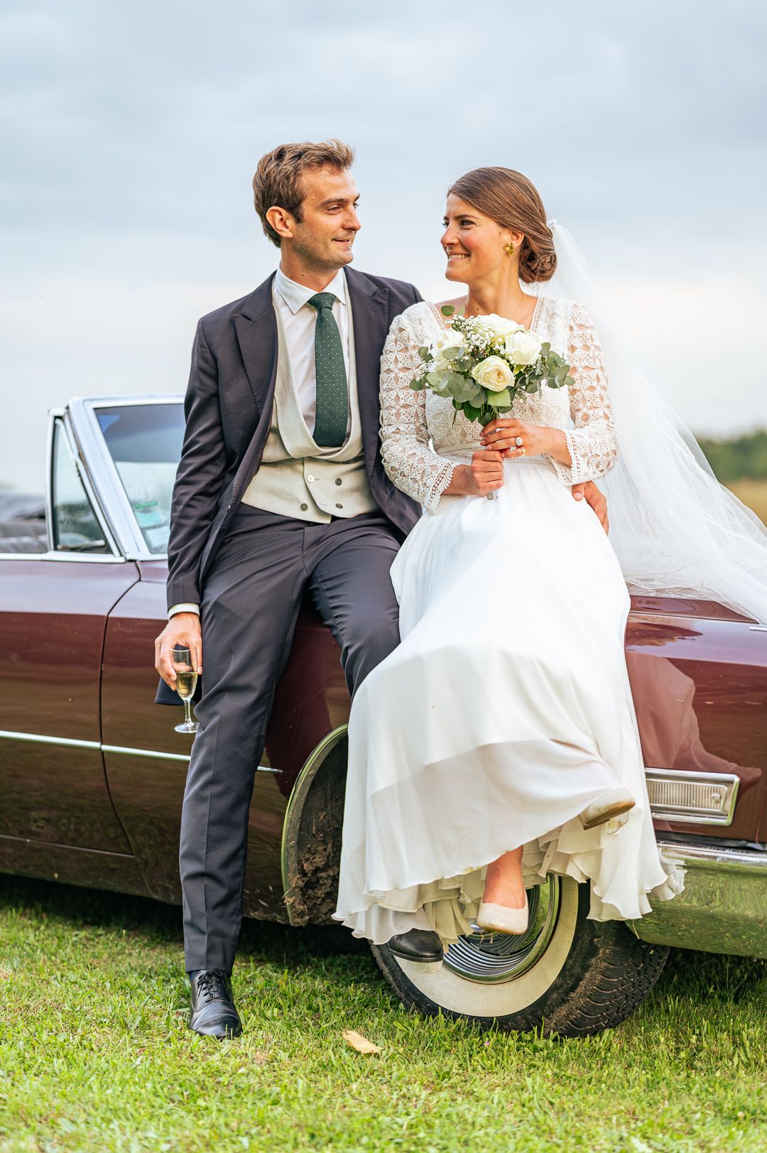 A newly married couple sits on a vintage car, sharing a joyful moment together.