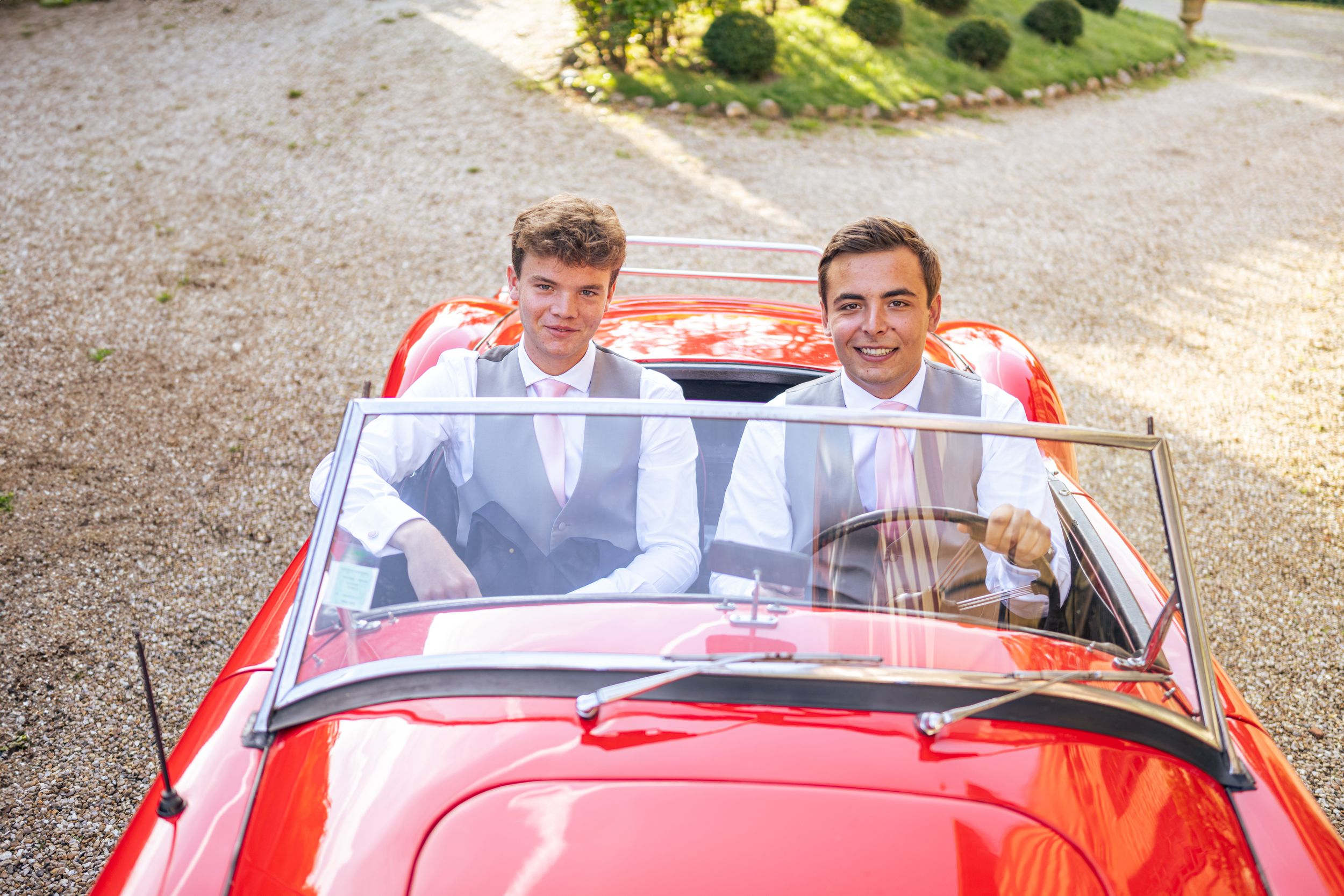 Two people smile while sitting in a bright red classic convertible car.