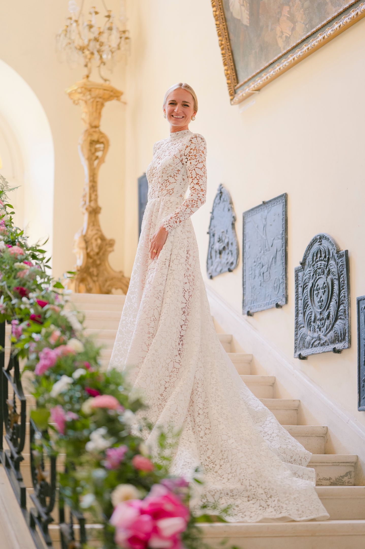 A bride in a detailed lace gown stands gracefully on a grand staircase with floral decorations.