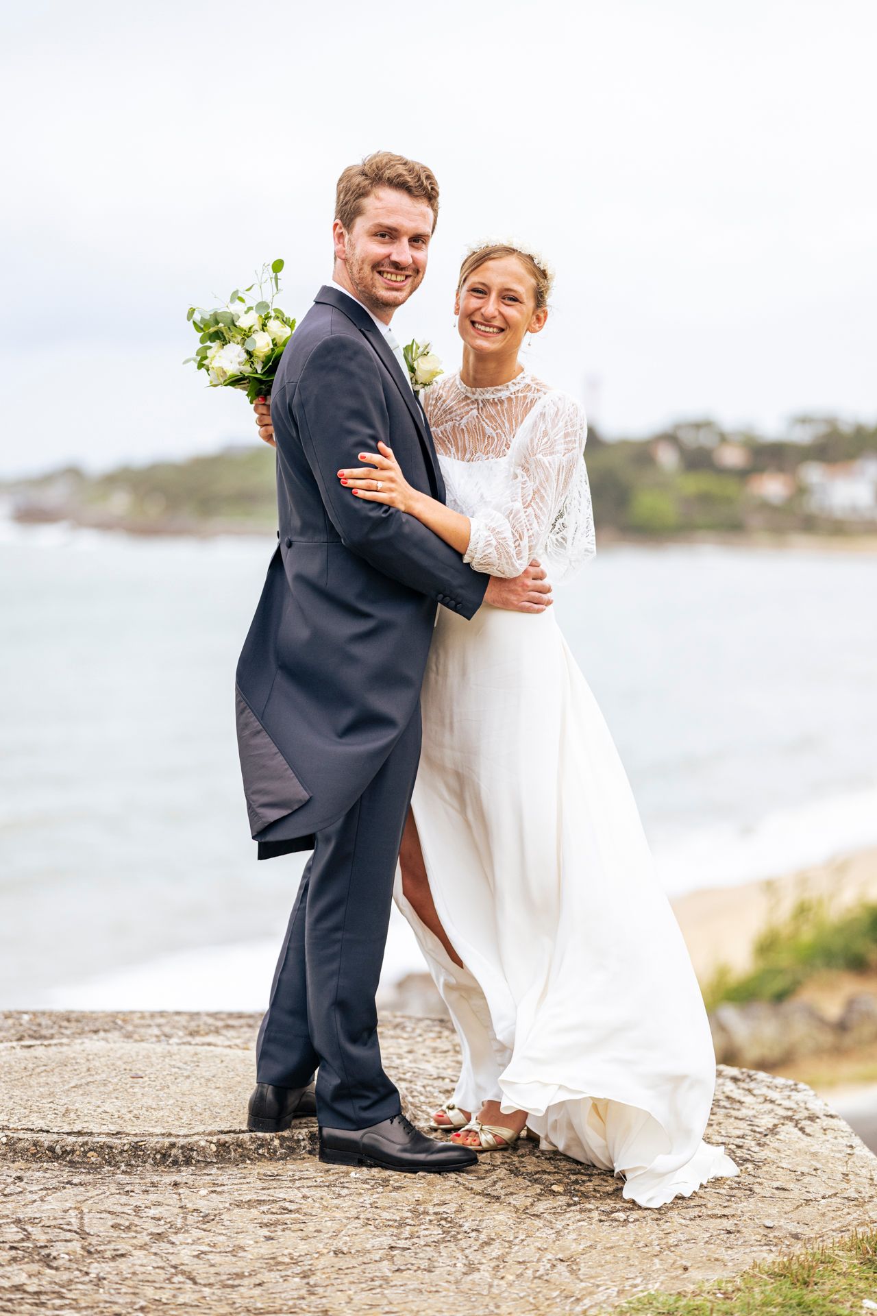 A newlywed couple posing by the ocean with the bride holding a white bouquet.