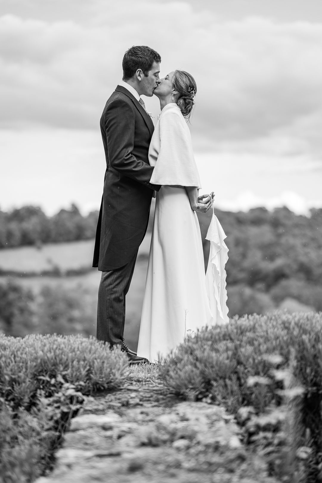 A black and white portrait of a wedding couple embracing on a hilltop path.