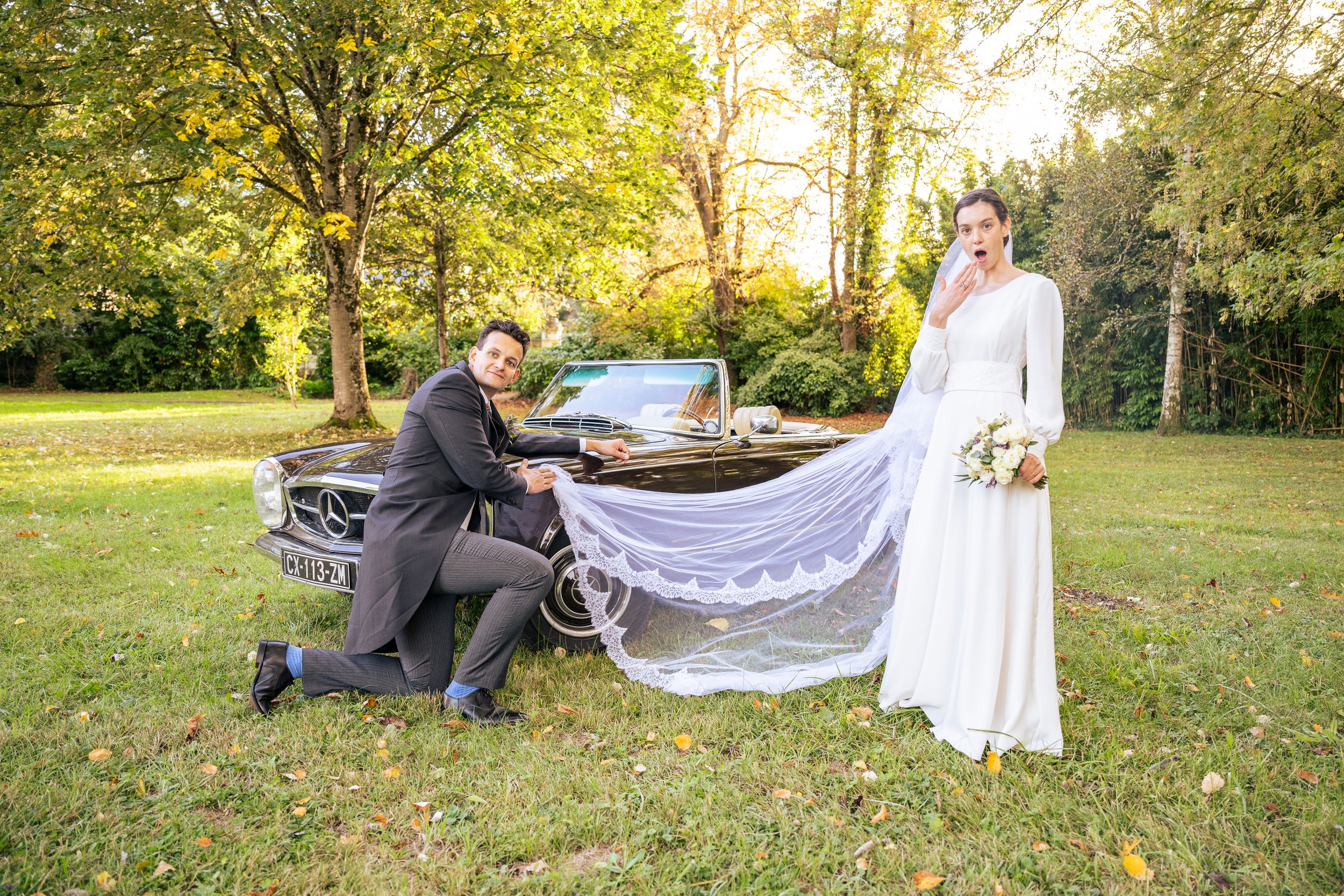 A wedding photographer arranges a bride's long flowing veil in a sunny garden setting.